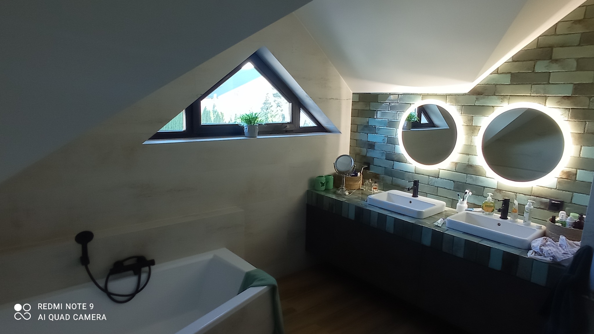Nowoczesna łazienka na poddaszu z dwoma umywalkami, okrągłymi lustrami z podświetleniem LED, wanną i trójkątnym oknem z widokiem na zieleń.