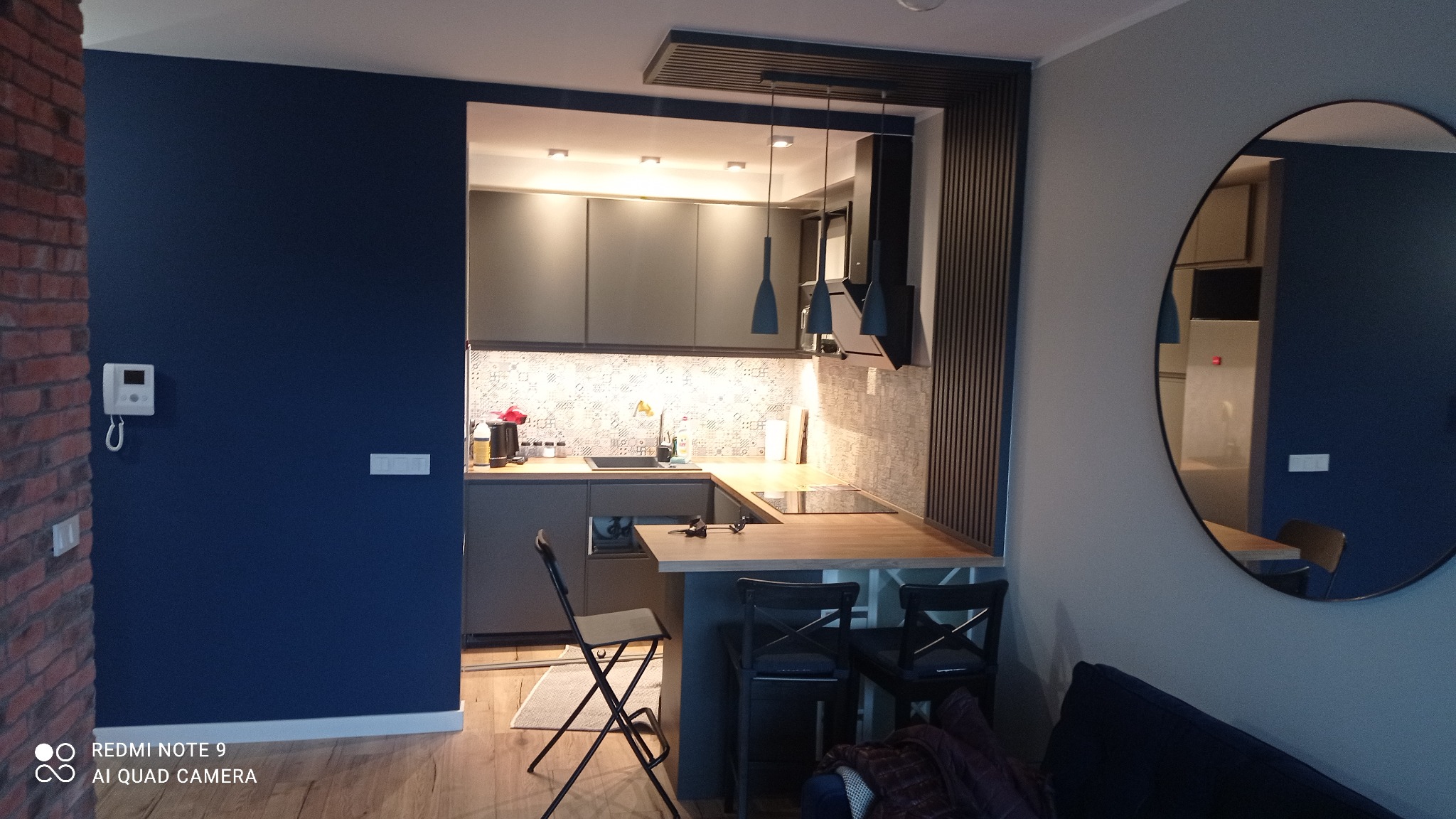 Nowoczesna kuchnia w odcieniach szarości i granatu z drewnianym blatem, widok z salonu, krzesła barowe, okrągłe lustro na ścianie