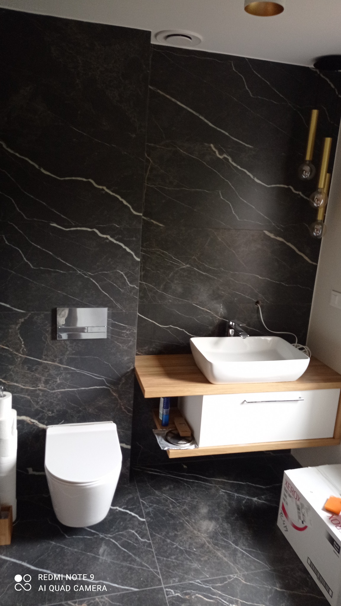 Nowoczesna łazienka z czarnymi płytkami imitującymi marmur, wisząca toaleta, umywalka nablatowa na drewnianym blacie i złote lampy wiszące.