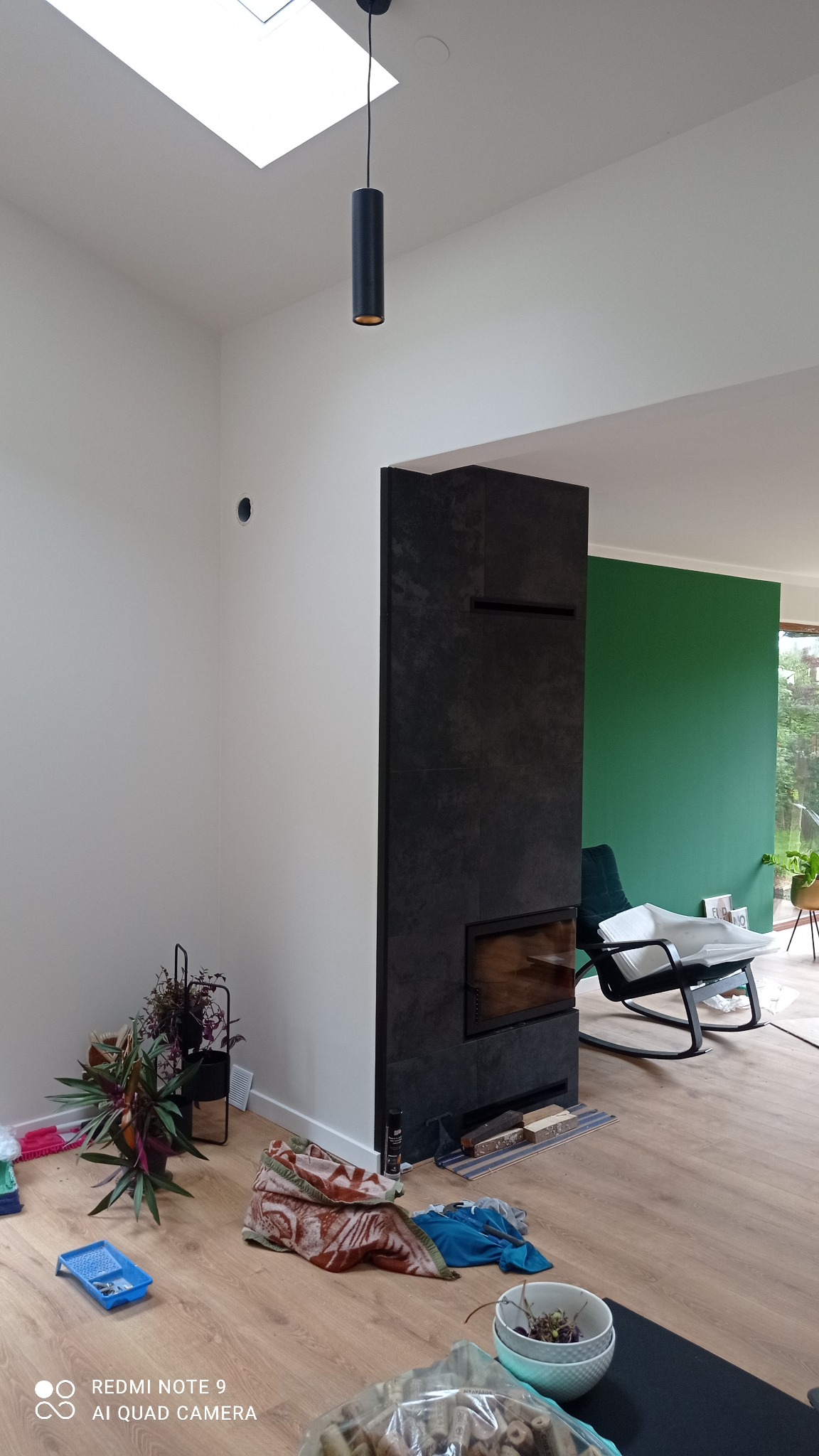 Wnętrze w trakcie prac wykończeniowych: białe ściany, czarny kominek z wkładem, sufit z oknem dachowym i wisząca lampa, podłoga wyłożona panelami, widoczne narzędzia i materiały budowlane, zielona...