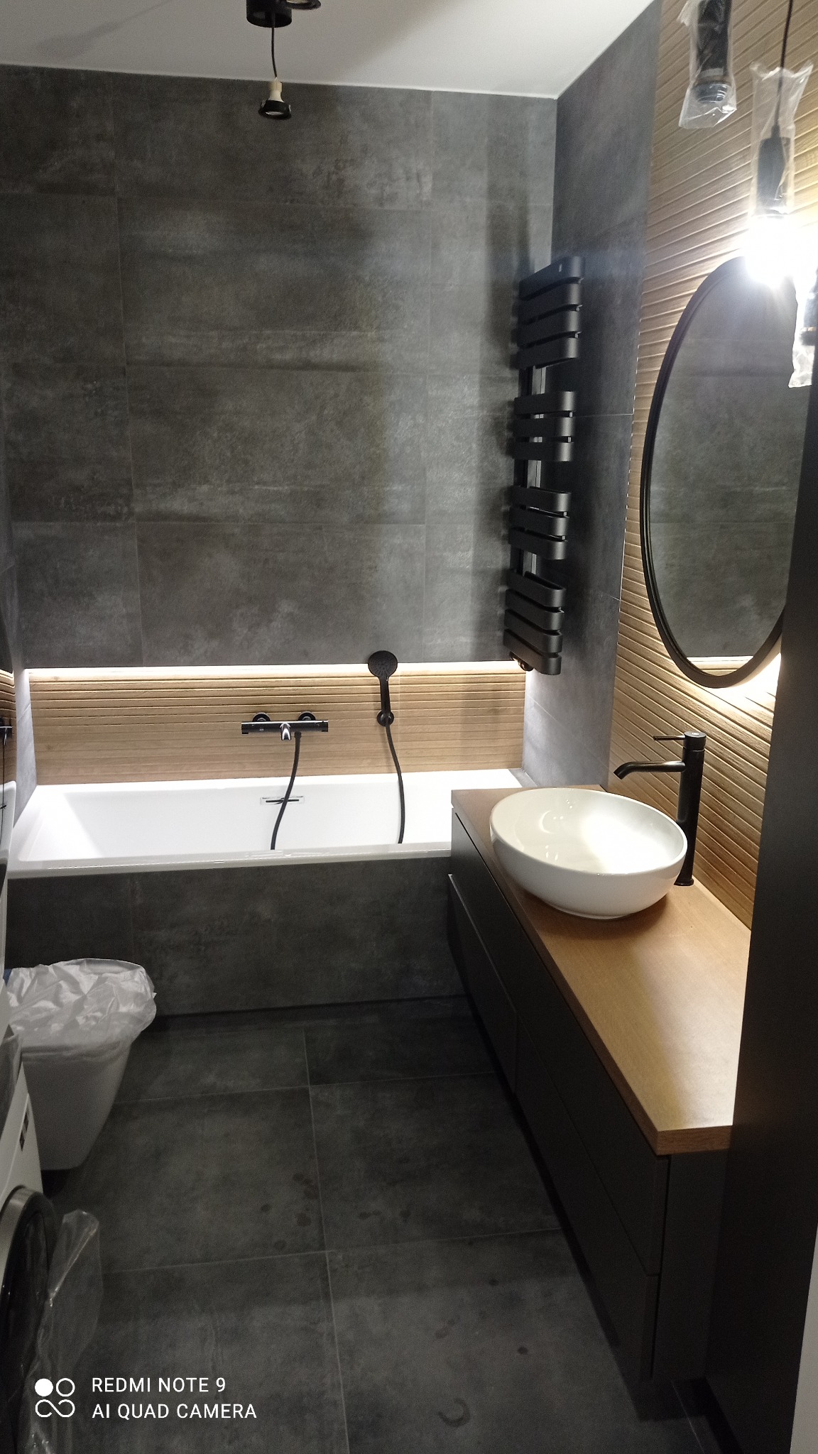 Nowoczesna łazienka z szarymi płytkami, drewnianymi akcentami i czarną armaturą; widoczna wanna, umywalka nablatowa i grzejnik drabinkowy.