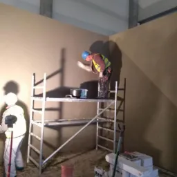 Pracownik w kasku i kamizelce na rusztowaniu wygładza ścianę w rogu pomieszczenia, drugi pracownik oświetla ścianę lampą, widoczne materiały budowlane i narzędzia.