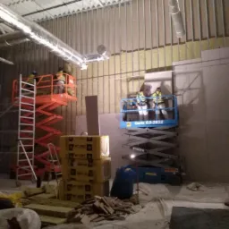 Prace wykończeniowe w dużym pomieszczeniu: pracownicy na podnośnikach montują izolację na metalowym stelażu ściany, widoczne materiały izolacyjne w żółtych opakowaniach i resztki materiałów...