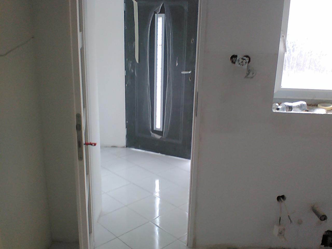 Wnętrze w trakcie remontu: widoczne białe płytki na podłodze, częściowo otwarte białe drzwi wewnętrzne z czerwoną klamką, szara drzwi wejściowe z długim oknem, niezabezpieczone gniazdka elektryczne...