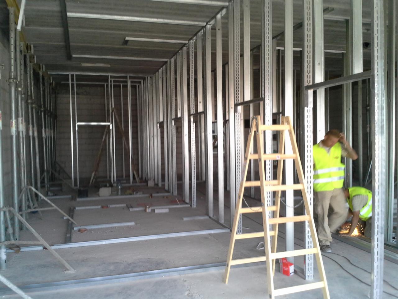 Wnętrze w trakcie budowy z widoczną metalową konstrukcją szkieletową ścian działowych, drewnianą drabiną na pierwszym planie i dwoma pracownikami w kamizelkach odblaskowych, z których jeden spawa...