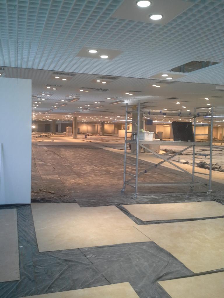 Rozległe wnętrze w trakcie prac wykończeniowych, widoczne rusztowanie, zabezpieczona podłoga i sufit kasetonowy z oświetleniem punktowym.
