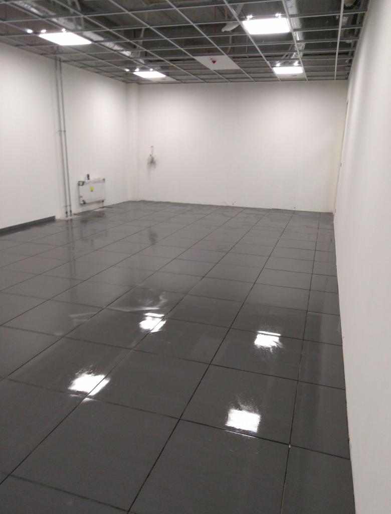 Puste pomieszczenie z szarą, błyszczącą podłogą wyłożoną kwadratowymi płytkami, białe ściany, widoczna instalacja pod sufitem rastrowym z oświetleniem.