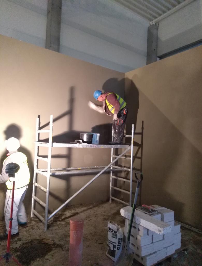 Pracownik w kasku i kamizelce na rusztowaniu wygładza ścianę w rogu pomieszczenia, drugi pracownik oświetla ścianę lampą, widoczne materiały budowlane i narzędzia.