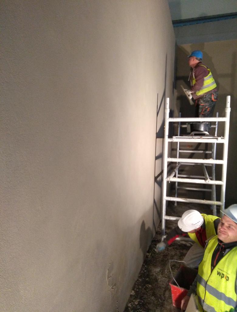 Dwóch pracowników w odblaskowych kamizelkach i kaskach szpachluje ścianę, jeden na drabinie, drugi z dołu, widoczny fragment ściany przed i po szpachlowaniu.