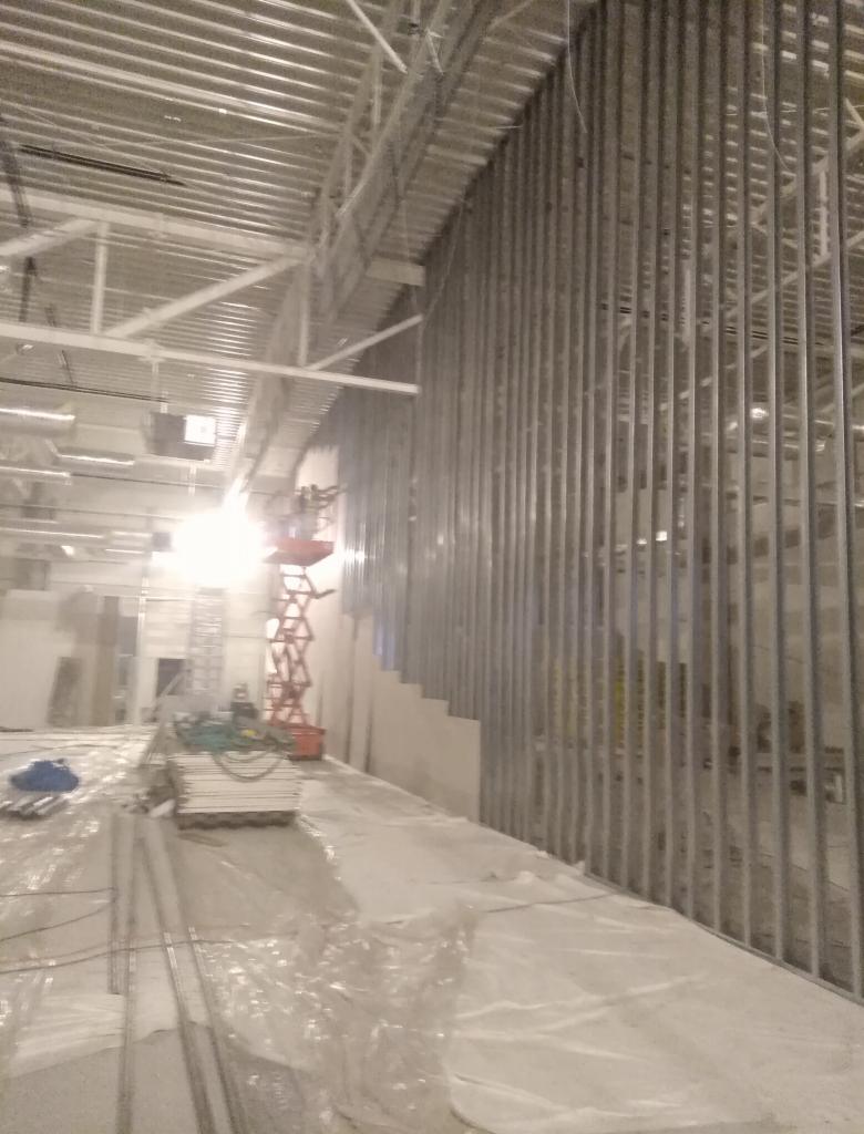 Montaż metalowego stelażu pod płytę karton-gips w dużym pomieszczeniu, widoczna podnośnik nożycowy i zabezpieczona folią podłoga.