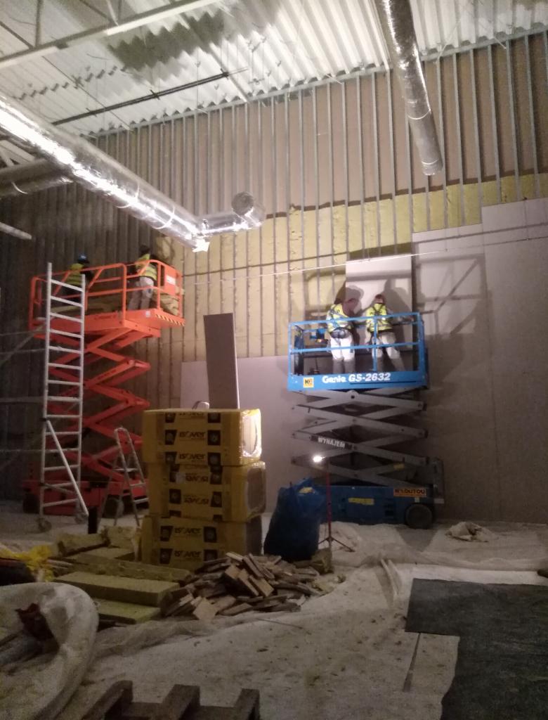 Prace wykończeniowe w dużym pomieszczeniu: pracownicy na podnośnikach montują izolację na metalowym stelażu ściany, widoczne materiały izolacyjne w żółtych opakowaniach i resztki materiałów...