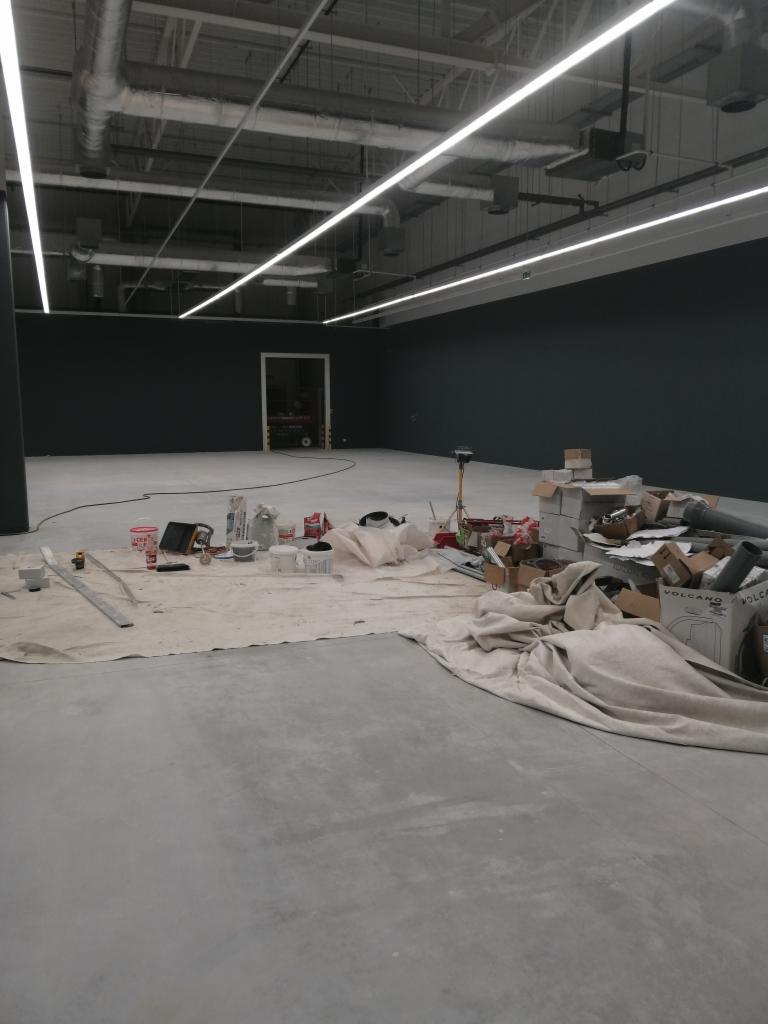 Wnętrze hali przemysłowej w trakcie prac wykończeniowych, widoczne rozłożone folie ochronne, narzędzia malarskie, puszki z farbą i kartony z materiałami budowlanymi.