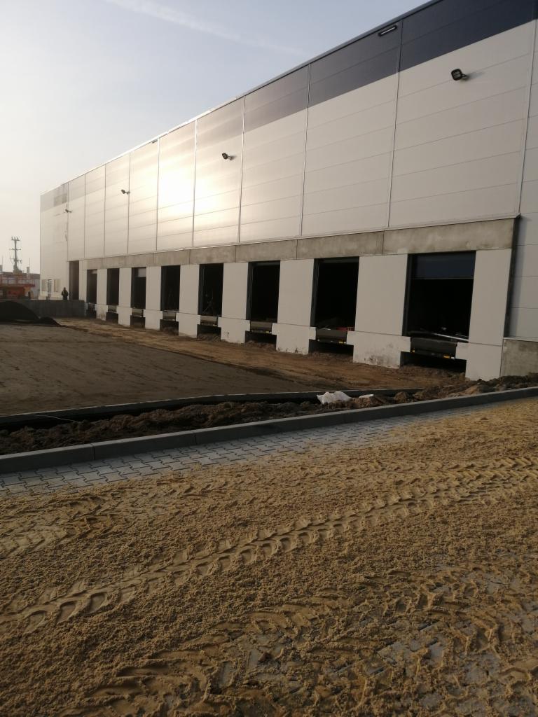 Budynek magazynowy w trakcie budowy, widoczne otwory okienne z przygotowanymi podmurówkami, teren wokół budynku z ziemią i śladami opon.