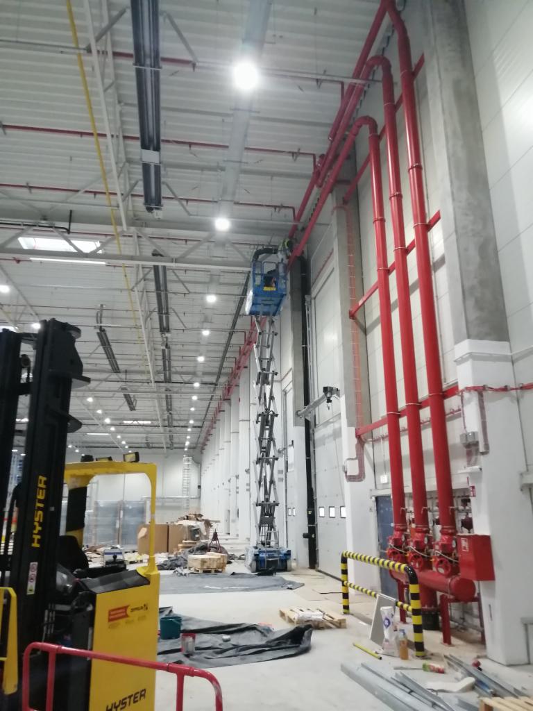 Montaż instalacji przeciwpożarowej w hali magazynowej z użyciem podnośnika nożycowego, widoczne czerwone rury i zawory, w tle regały i materiały budowlane.