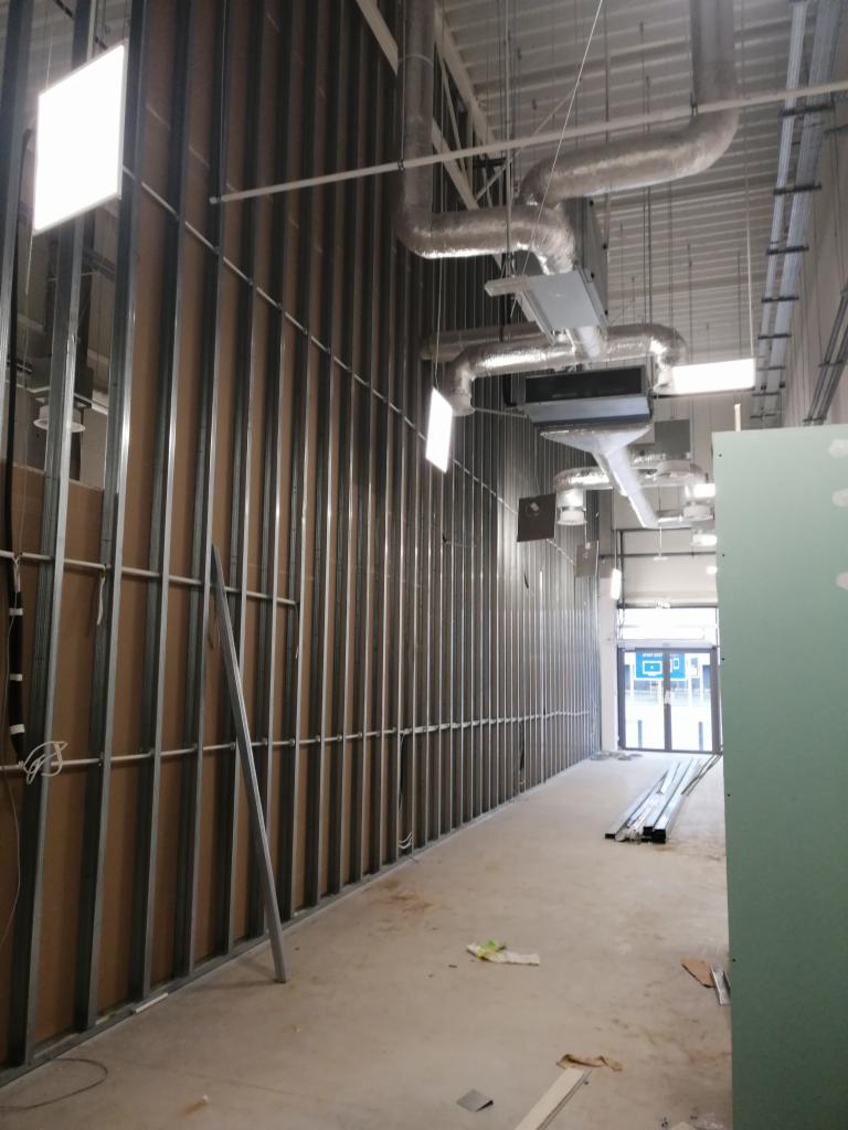 Wnętrze w trakcie budowy: metalowy szkielet ściany, widoczne instalacje wentylacyjne pod sufitem, surowy beton na podłodze, przygotowane profile leżące na podłodze, w tle przeszklone drzwi wejściowe.