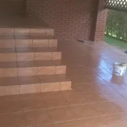 Wyłożone brązowymi płytkami schody zewnętrzne i taras, widoczny kubeł po farbie i narzędzia, ceglana ściana w tle.