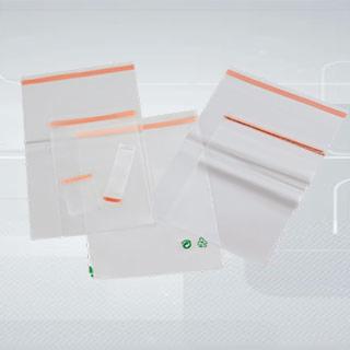 Zestaw przezroczystych torebek strunowych z pomarańczowym paskiem zamykającym, ułożonych pod różnymi kątami na jasnym tle z subtelnym wzorem. Widoczne małe zielone symbole recyklingu na jednej...