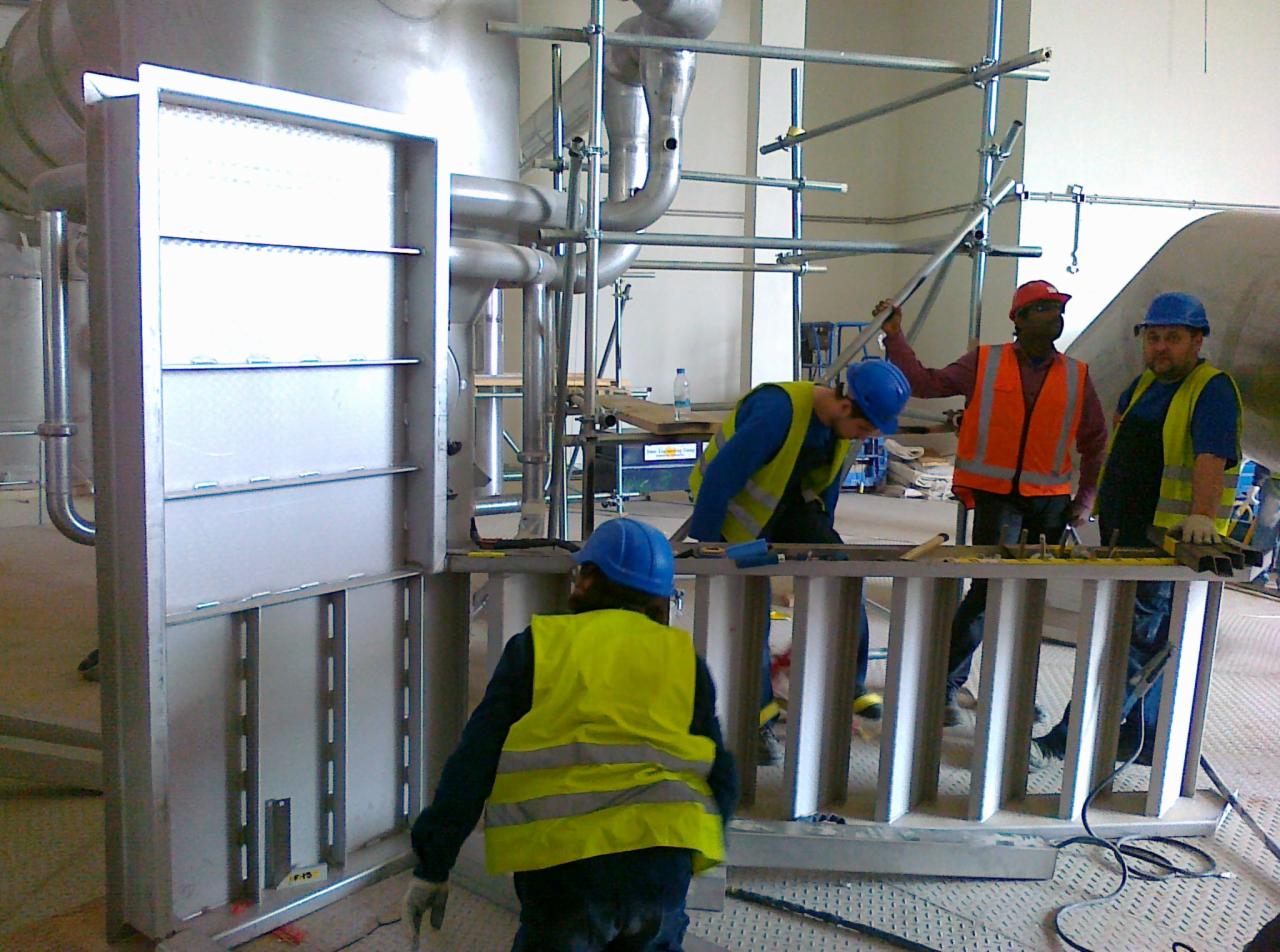 Montaż konstrukcji stalowej w hali przemysłowej w Krakowie, widoczni pracownicy w kaskach i kamizelkach odblaskowych, rusztowania i elementy instalacji w tle.