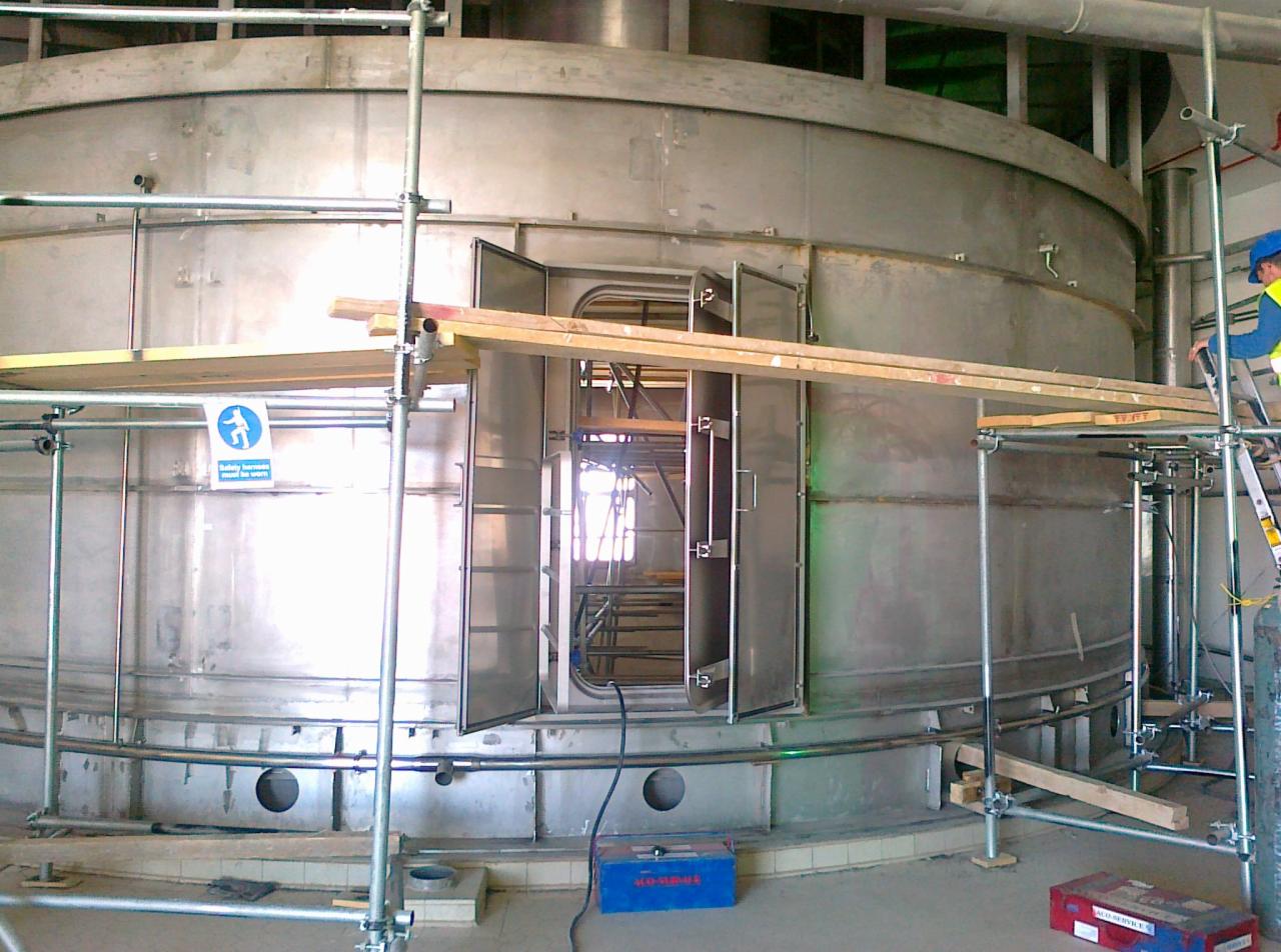 Wielkogabarytowy, metalowy zbiornik cylindryczny z otwartymi drzwiami inspekcyjnymi, otoczony rusztowaniem budowlanym i z pracownikiem w kasku na drabinie.