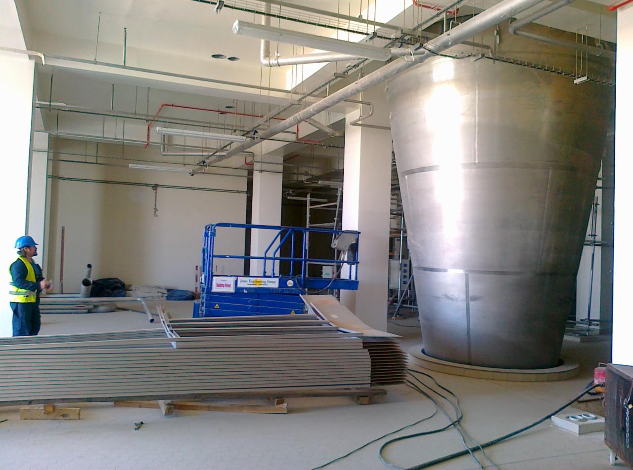 Wnętrze hali w trakcie budowy z widocznym pracownikiem w kasku, podnośnikiem nożycowym i elementami konstrukcji stalowej w tle.