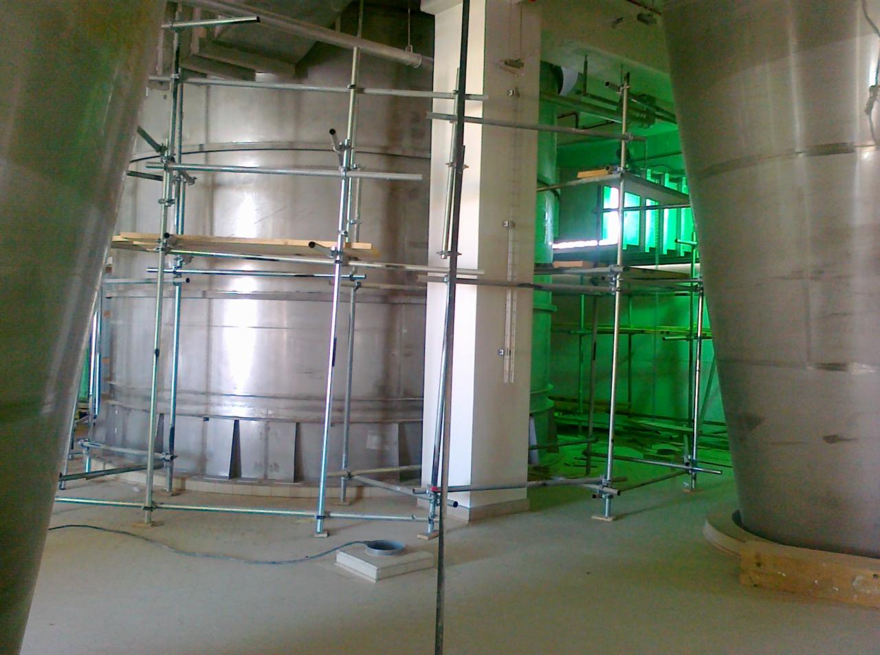 Wnętrze hali przemysłowej w trakcie budowy, widoczne metalowe zbiorniki otoczone rusztowaniami, z zielonymi elementami w tle.