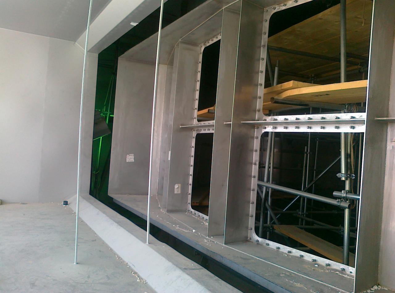 Wnętrze budowanego obiektu z widoczną stalową konstrukcją szkieletową z otworami, mocowaną za pomocą śrub, z rusztowaniem w tle.