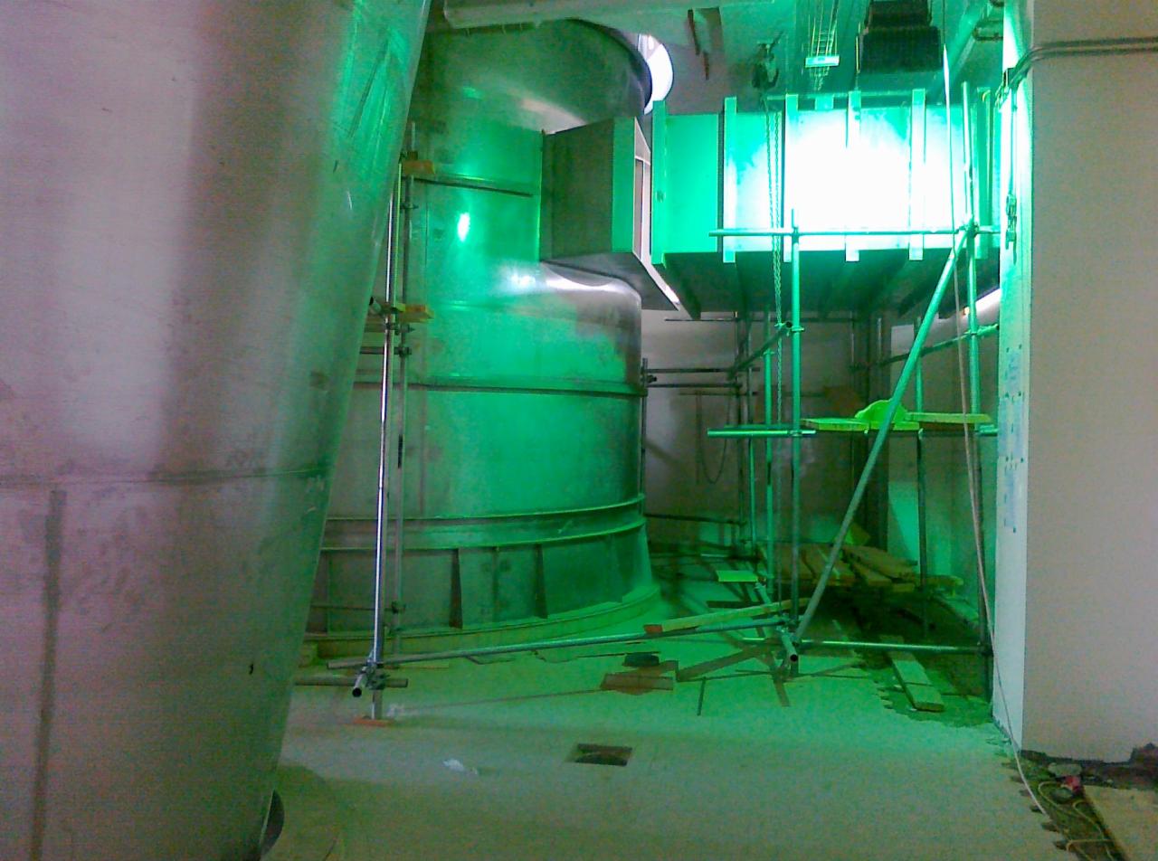 Wnętrze hali w trakcie budowy, widoczne elementy konstrukcji stalowej, rusztowania i system wentylacji, oświetlone zielonym światłem.