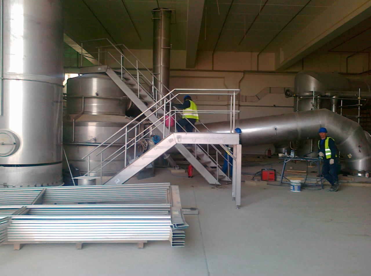 Wnętrze hali produkcyjnej z dużymi, stalowymi zbiornikami, rurami i schodami. Dwóch pracowników w odblaskowych kamizelkach i kaskach, jeden wchodzi po schodach, drugi stoi obok spawarki.