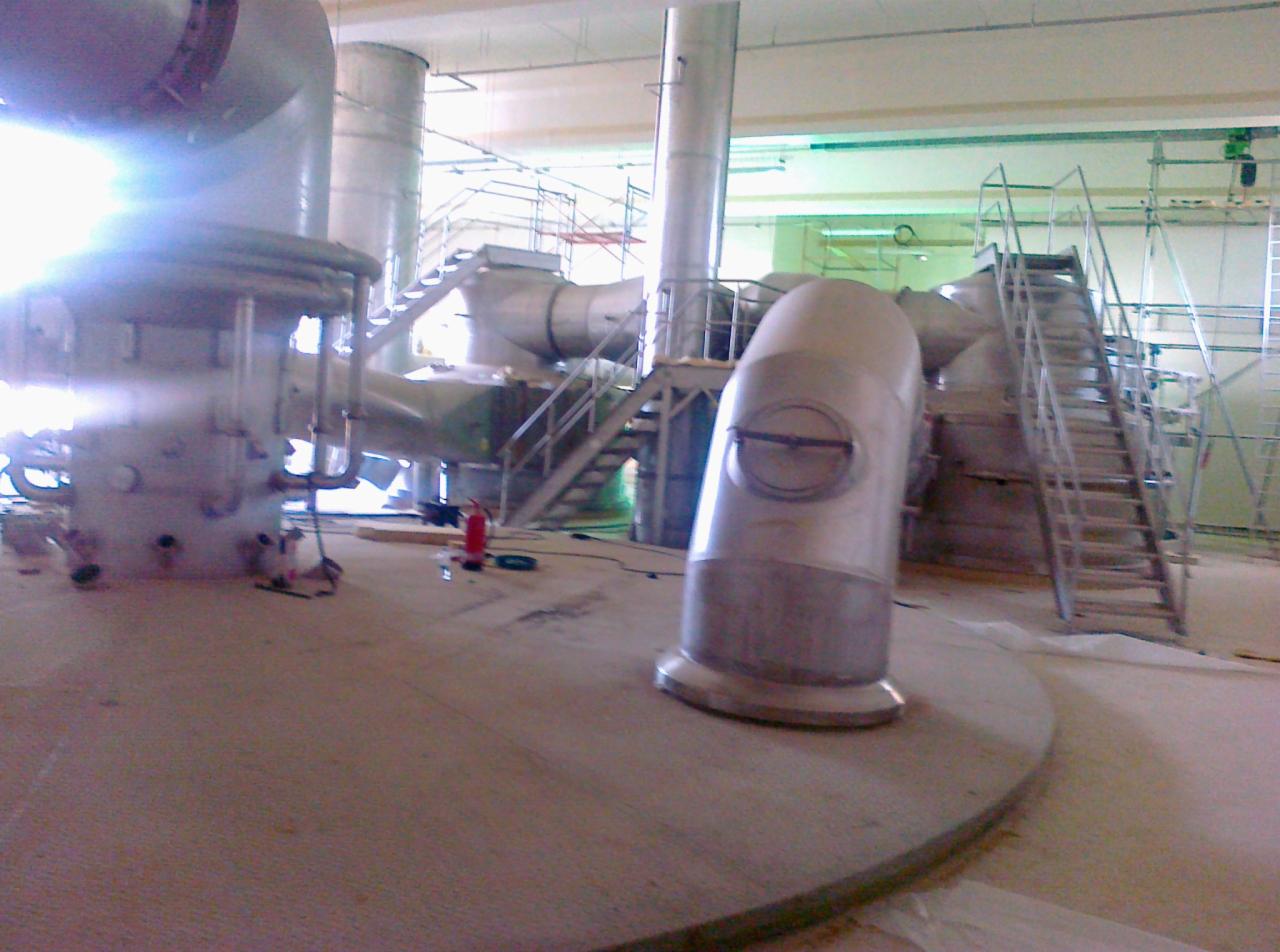 Wnętrze hali w trakcie budowy: widoczne metalowe rury, konstrukcje schodów i rusztowania, czerwona gaśnica na podłodze.