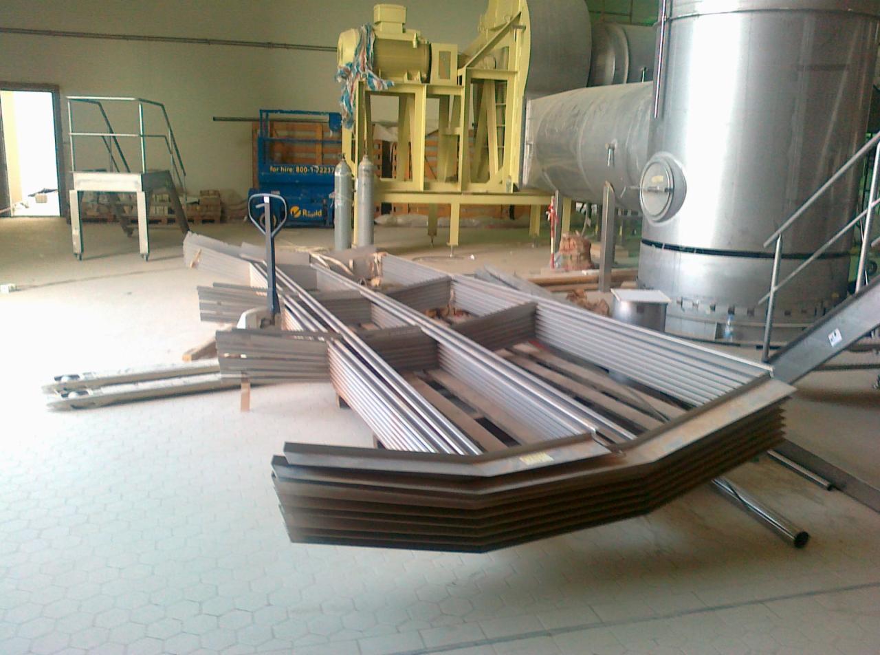 Stalowe elementy konstrukcyjne, prawdopodobnie fragmenty ramy, ułożone na palecie transportowej w hali, w tle widoczny zbiornik i podnośnik.