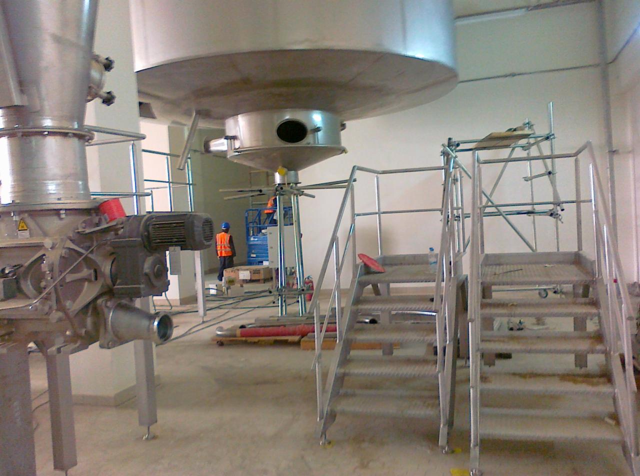 Wnętrze hali produkcyjnej w trakcie montażu dużych, metalowych zbiorników i instalacji, widoczne schody z metalową konstrukcją oraz pracownik w odblaskowej kamizelce na podnośniku.