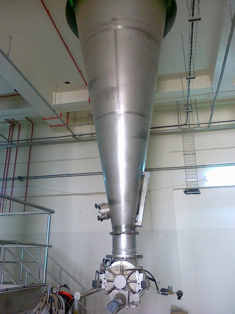 Wysoki, stożkowy zbiornik ze stali nierdzewnej z zamontowanym na dole dozownikiem obrotowym w hali przemysłowej, widoczne rury i kable pod sufitem.