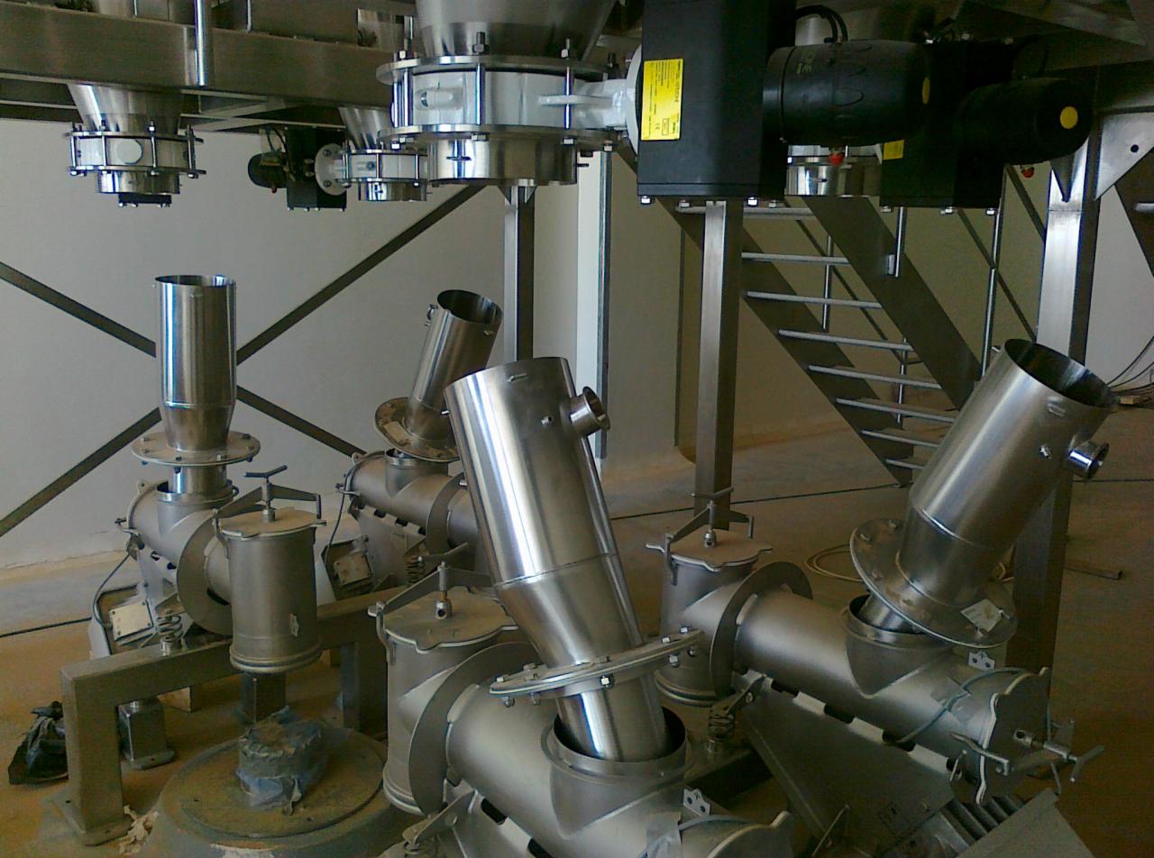 Kompleksowa instalacja przemysłowa z polerowanej stali nierdzewnej, składająca się z pionowych i skośnych rur, zbiorników oraz elementów mocujących na tle białej ściany i schodów.