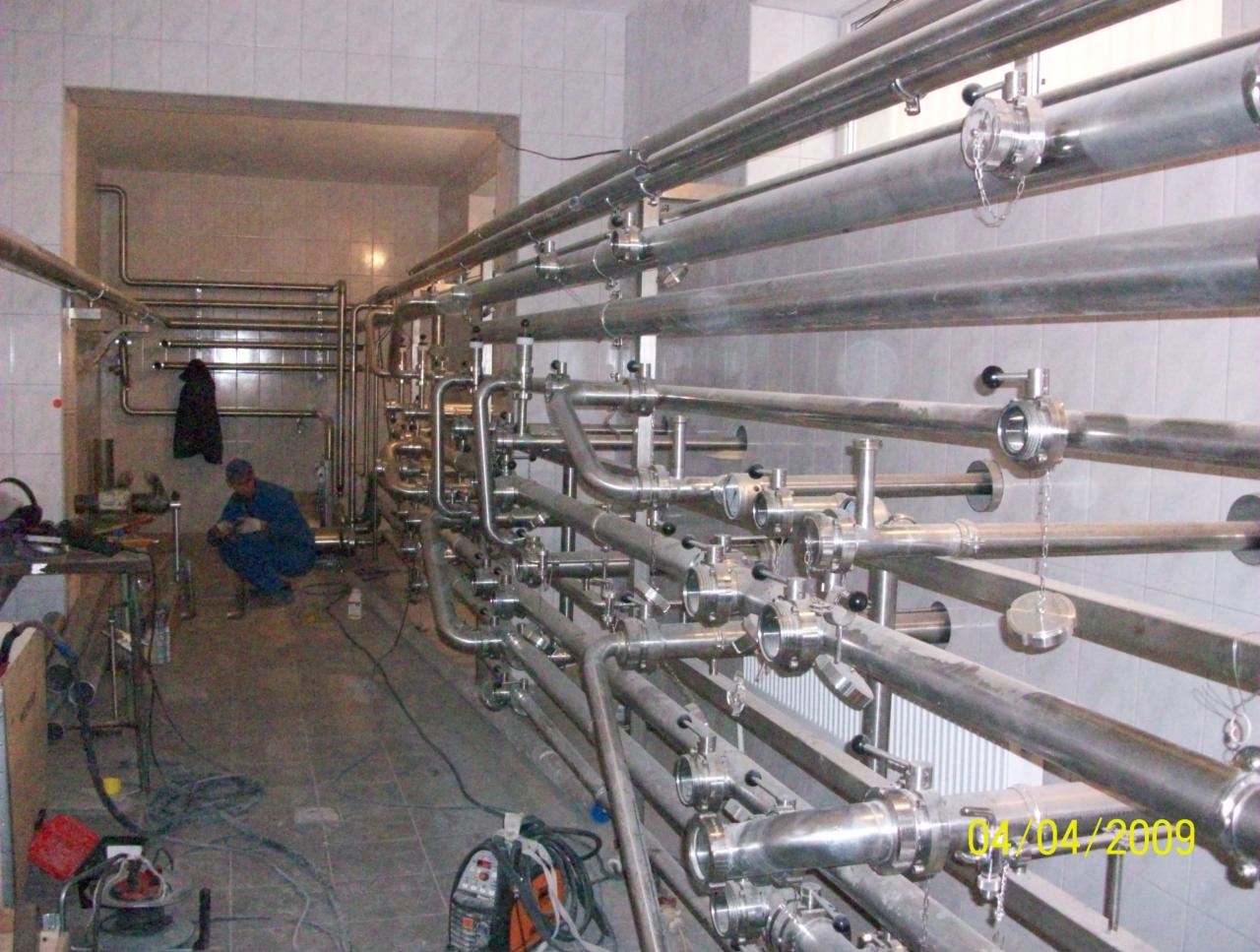 Instalacja rurociągów ze stali nierdzewnej w pomieszczeniu wyłożonym kafelkami, widoczny pracownik w kombinezonie i sprzęt spawalniczy.