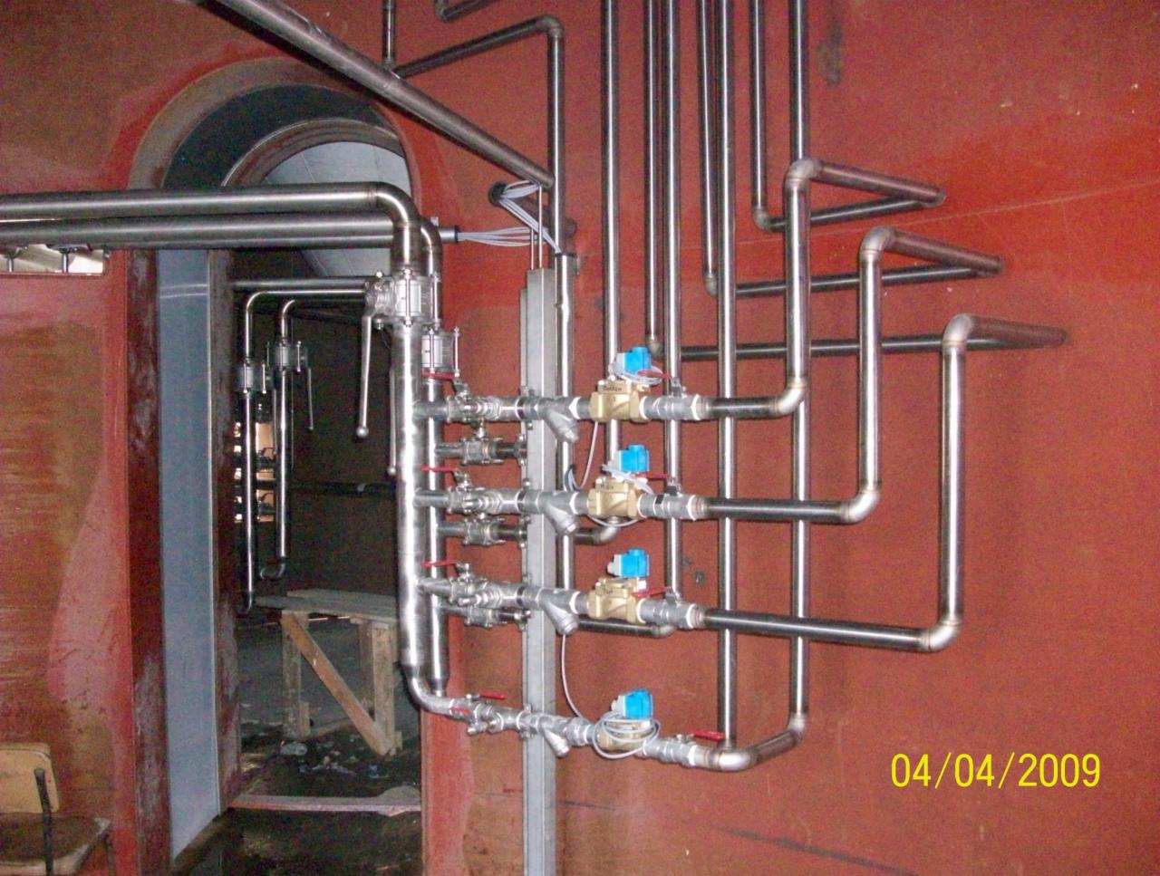 Instalacja rurociągów ze stali nierdzewnej z zaworami kulowymi wewnątrz pomalowanego na czerwono zbiornika lub pomieszczenia, widoczne ślady korozji i zabrudzenia.