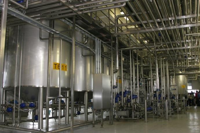 Wnętrze hali produkcyjnej z szeregiem stalowych zbiorników i rur, oznaczonych żółtymi etykietami, tworzących skomplikowany system instalacji.