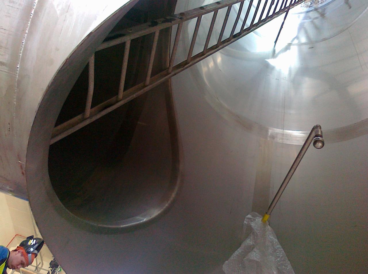 Wnętrze metalowego zbiornika z drabinką i spawaczem w kasku, widok z dołu w trakcie prac spawalniczych.