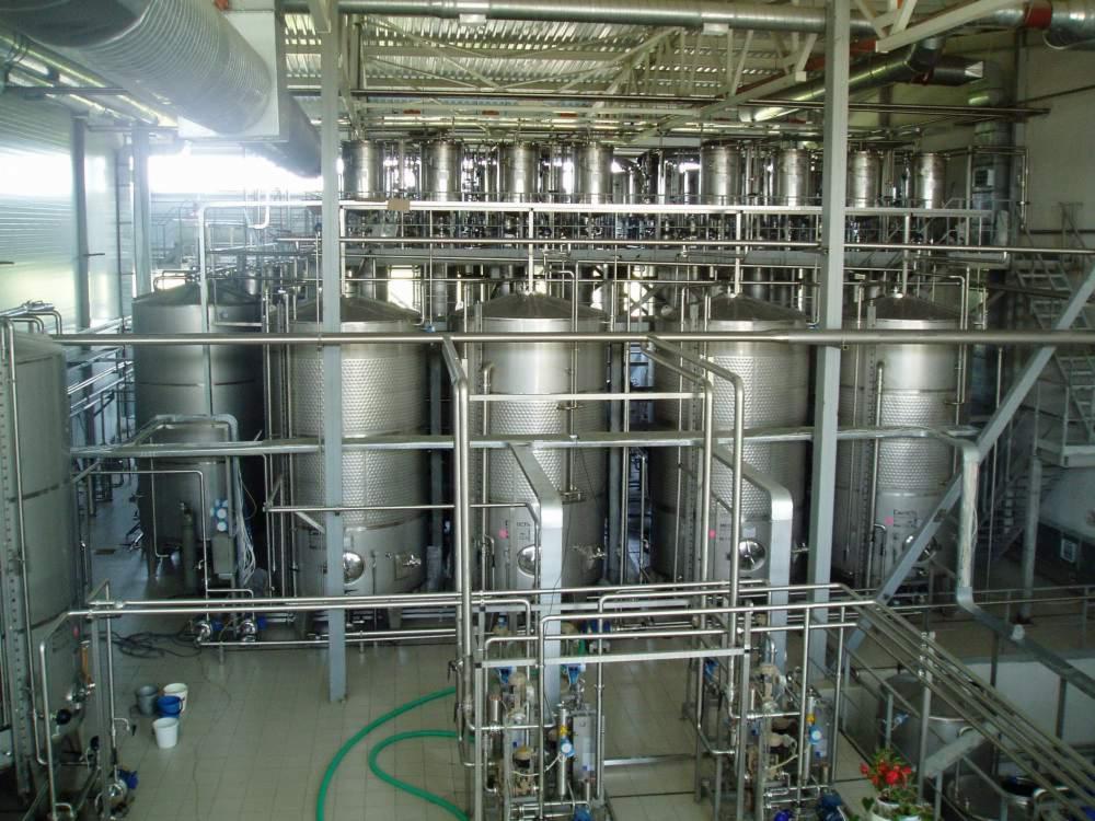 Wnętrze hali produkcyjnej z dużą ilością zbiorników ze stali nierdzewnej, rur i instalacji, widoczne schody i oświetlenie sufitowe.