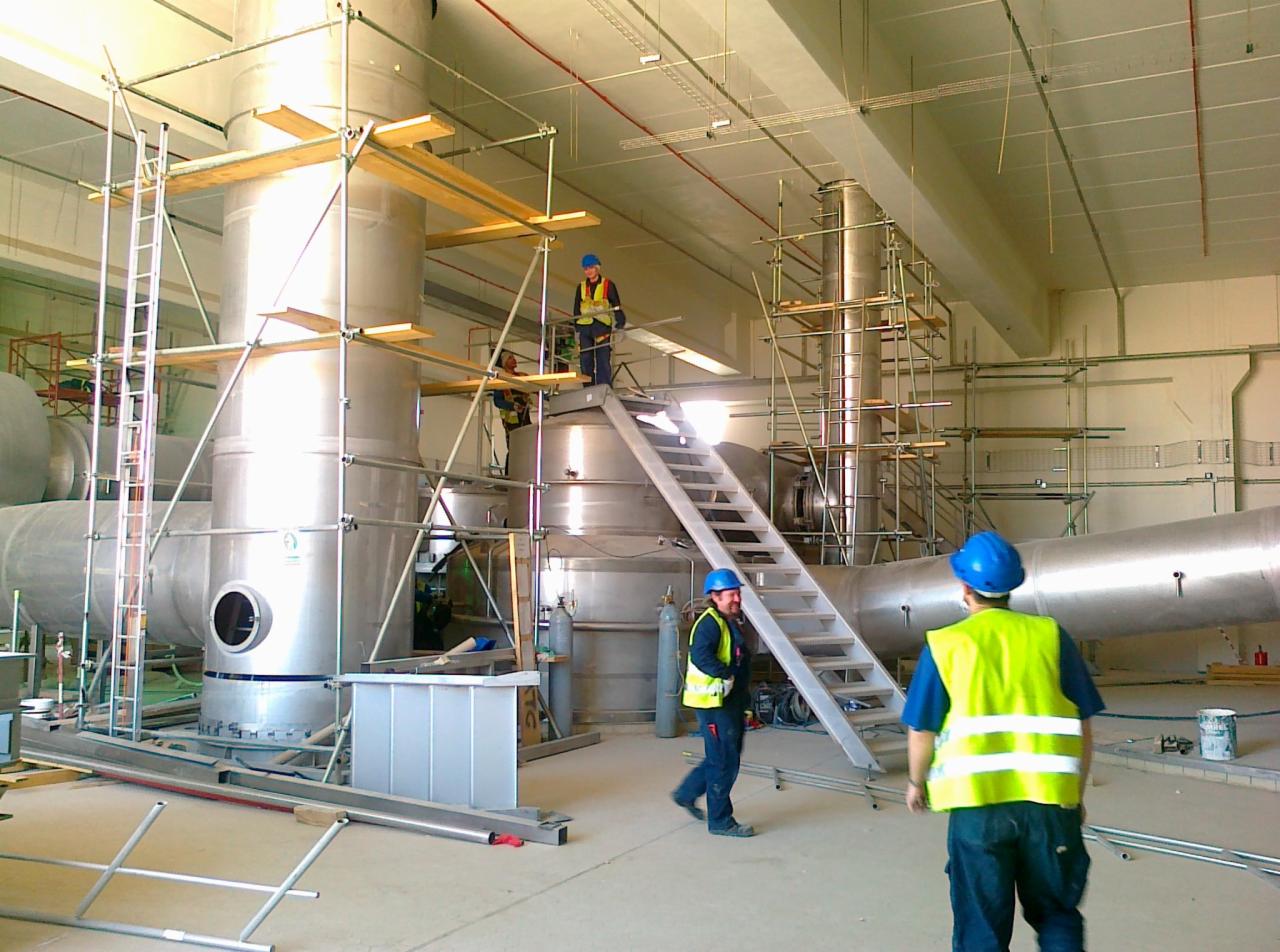 Montaż stalowej instalacji przemysłowej wewnątrz hali, widoczne rusztowania i pracownicy w kaskach i kamizelkach odblaskowych, jasne wnętrze.