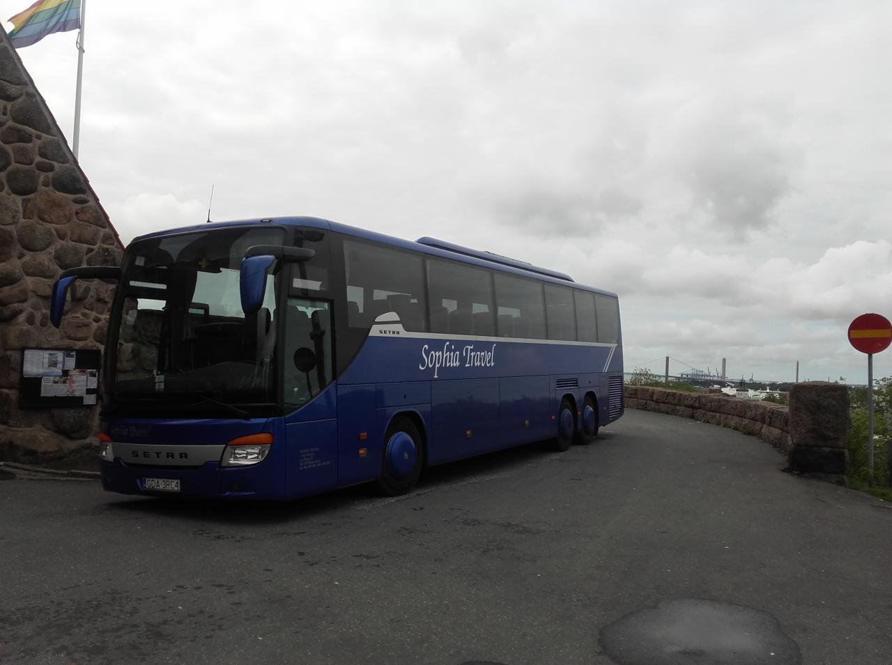 Niebieski autokar Setra 'Sophia Travel' zaparkowany na tle kamiennego muru i pochmurnego nieba, widoczny maszt z tęczową flagą.