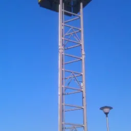 Wysoki, żółty pylon reklamowy z napisem 'STOP SHOP' na tle błękitnego nieba, widoczna metalowa konstrukcja wsporcza i lampa uliczna.