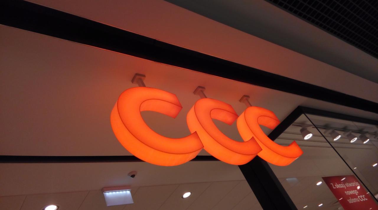 Podświetlane, pomarańczowe logo 'CCC' zamontowane pod sufitem w galerii handlowej, widoczne z dołu pod kątem.