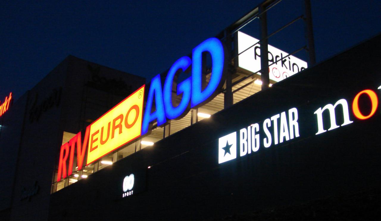 Oświetlone szyldy reklamowe sklepów RTV Euro AGD, Big Star i innych na elewacji budynku po zmroku.