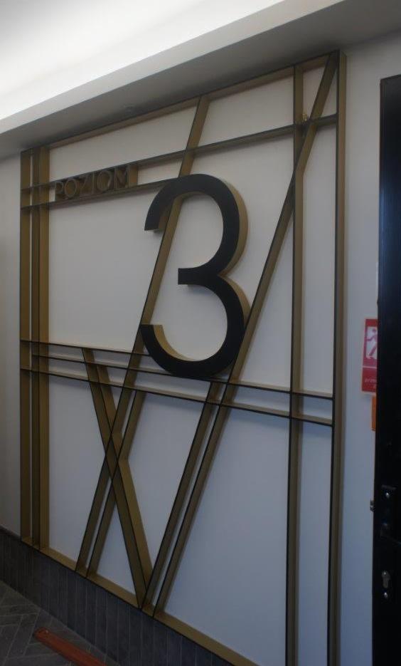 Numer '3' w czarnej obwódce, napis 'POZIOM' i dekoracyjne złote metalowe elementy na białej ścianie, wskazujące na numer piętra w budynku.