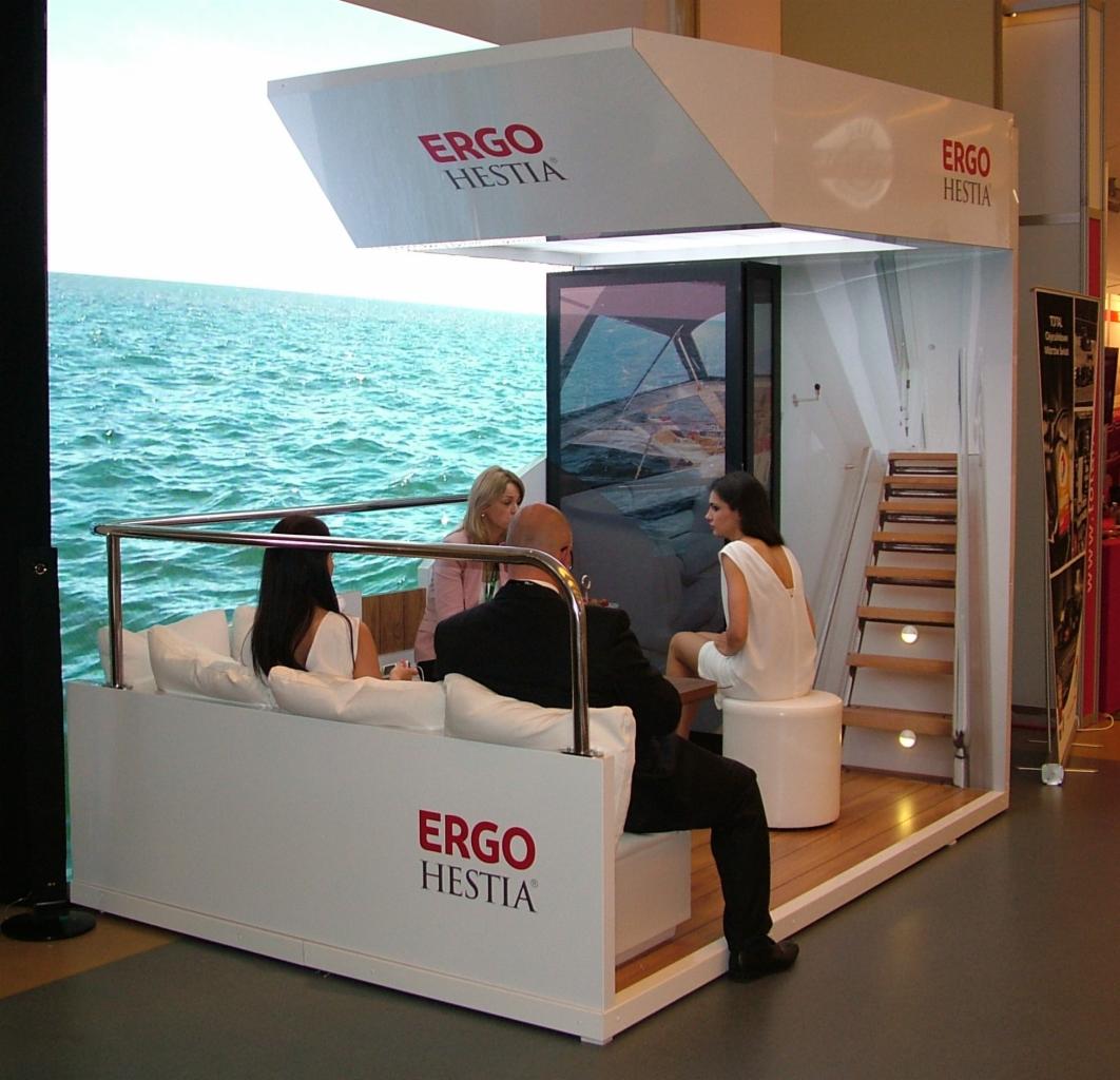 Stoisko targowe z wyświetlaczem LED prezentującym widok morza, logo firmy ERGO HESTIA na ścianach, oraz siedzącymi osobami.