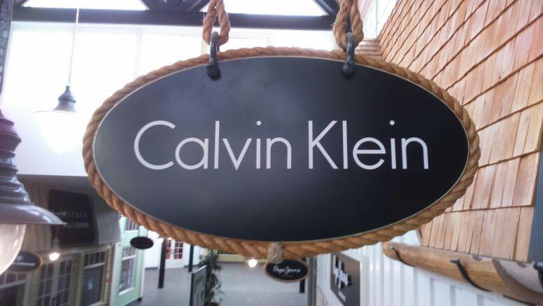 Owalny szyld z nazwą 'Calvin Klein' zawieszony na sznurze, z drewnianym tłem przypominającym dachówkę.