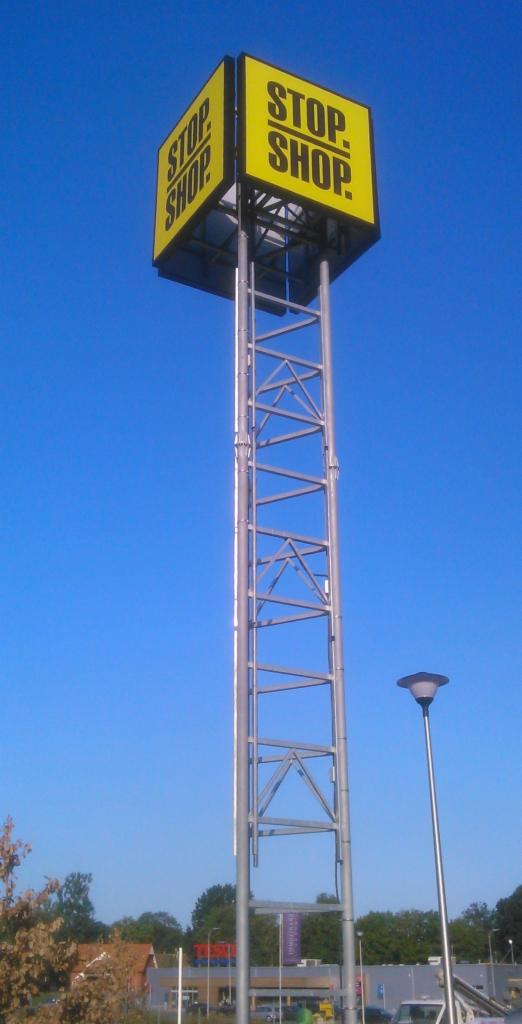 Wysoki, żółty pylon reklamowy z napisem 'STOP SHOP' na tle błękitnego nieba, widoczna metalowa konstrukcja wsporcza i lampa uliczna.