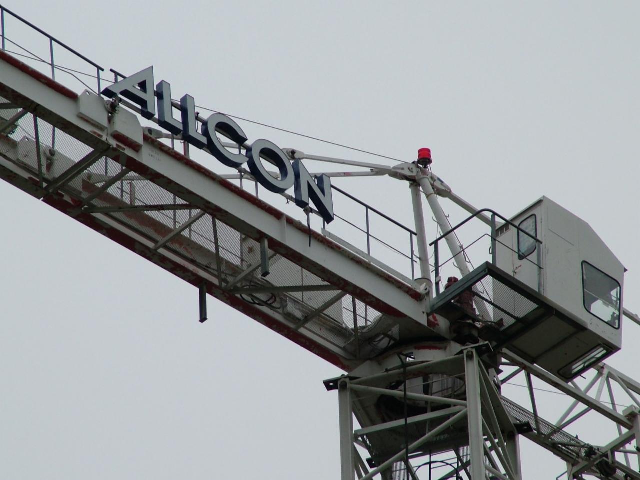 Biały dźwig budowlany z niebieskim napisem 'ALLCON' na ramieniu, widoczny pod kątem od dołu, na tle szarego nieba.