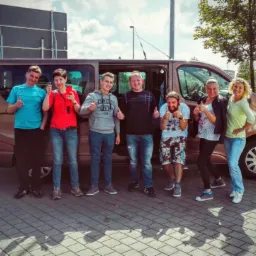 Grupa siedmiu osób, w tym jedna z brodą, pozuje z podniesionymi kciukami przed brązowym vanem z otwartymi drzwiami na parkingu z kostki brukowej, w tle stacja benzynowa i ciężarówka.