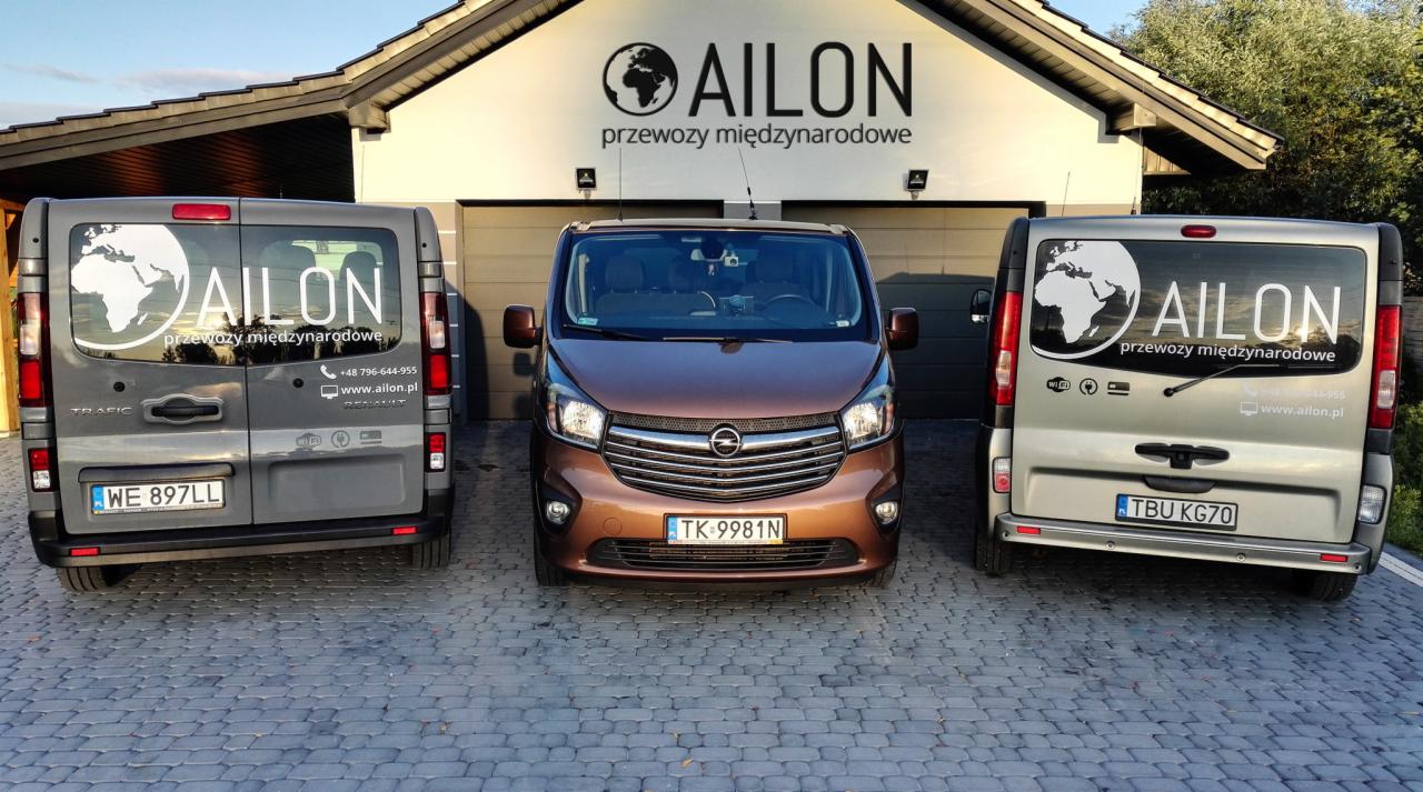 Trzy busy firmy Ailon przewozy międzynarodowe zaparkowane na brukowanym podjeździe przed budynkiem z logo firmy.